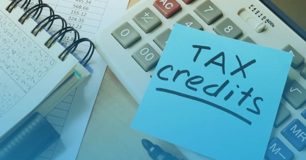 calculator, sheet of paper with numbers and a notebook in the background with a sticky note that says "Tax Credit"