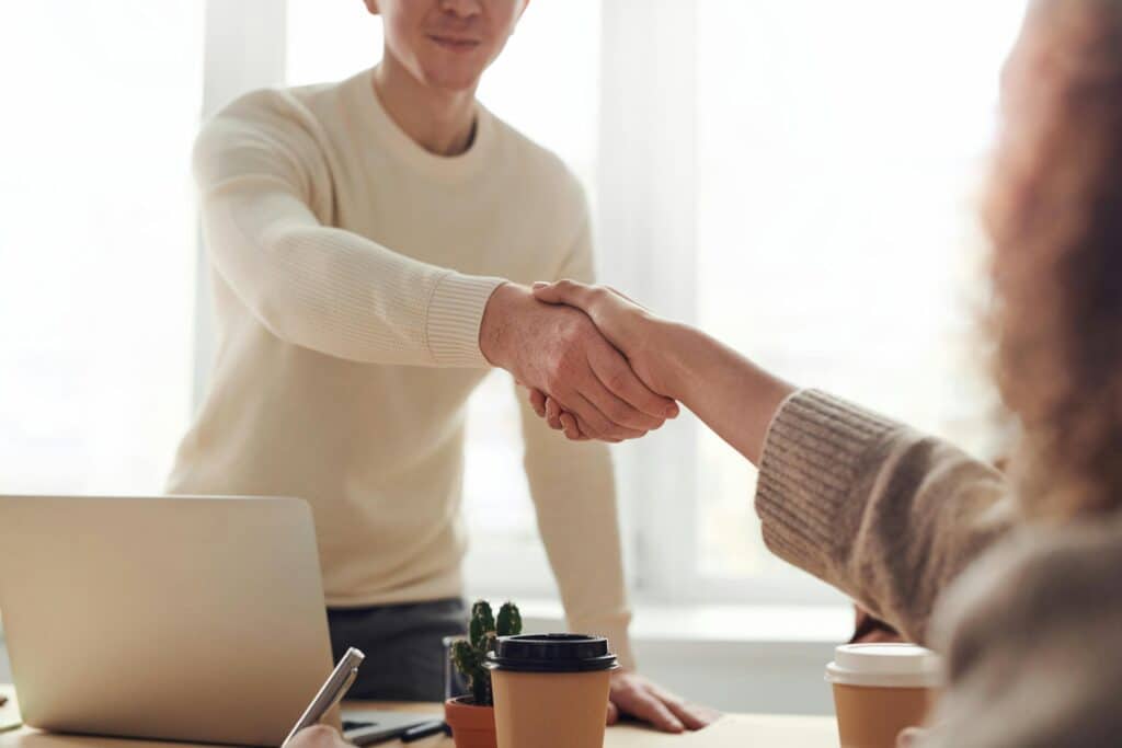 man in white shirt standing behind laptop shakes woman with curly hair's hand